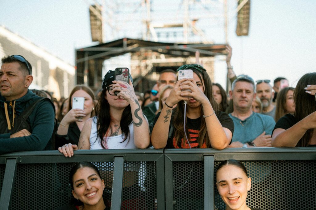 A lively crowd takes photos during an outdoor music concert on a sunny day.